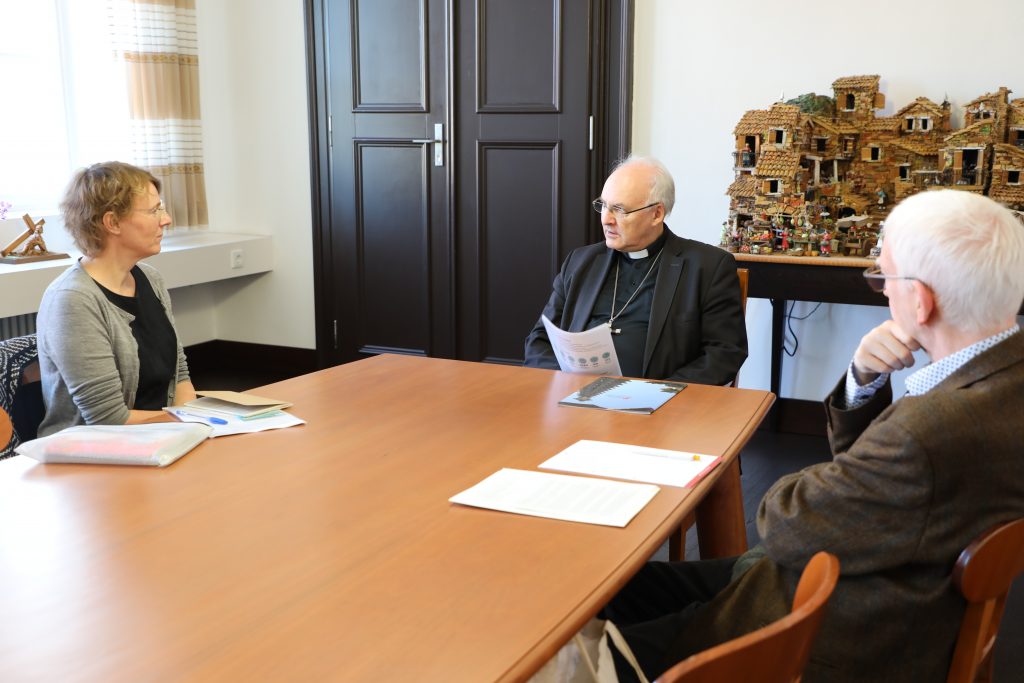 Drei Personen sitzen um einen Tisch in einem Büro und unterhalten sich über Migrationsfragen. Auf dem Tisch liegen Papiere und Notizbücher, im Hintergrund sind ein Figurenständer und eine Raphaelswerk-Anlaufstelle zu sehen.
