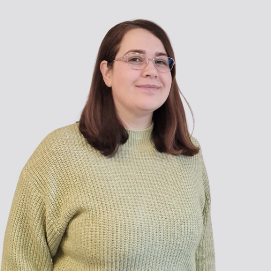 Eine Person mit mittellangem braunem Haar und Brille, die einen hellgr&uuml;nen Pullover tr&auml;gt, steht vor einem schlichten hellgrauen Hintergrund und l&auml;chelt leicht.