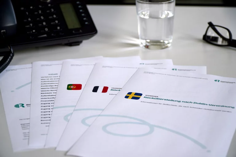 Dublin-Factsheets des Raphaelswerks mit verschiedenen Länderflaggen, ein Telefon, ein Glas Wasser und eine Brille auf einem Schreibtisch.