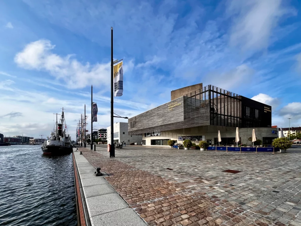 Modernes Gebäude am Wasser mit großen Fenstern und Sitzplätzen im Freien, direkt neben einem angedockten Schiff an einem sonnigen Tag mit teilweise bewölktem Himmel.