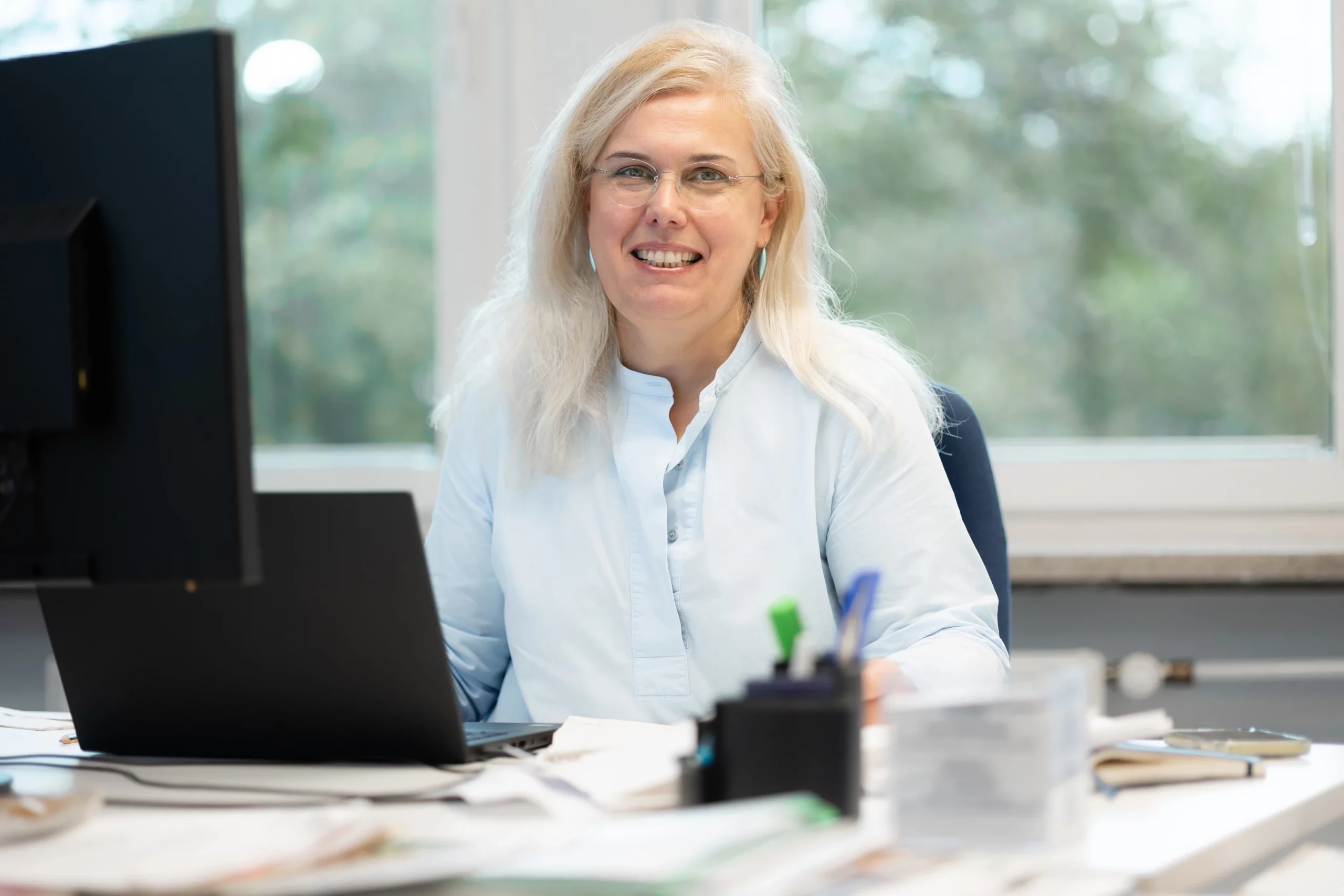 Eine Frau mit langen Haaren und Brille sitzt an einem Schreibtisch mit einem Computer, Papieren und Büromaterial und lächelt in die Kamera in einem gut beleuchteten Büro.