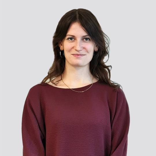 Laura Böllstorf, eine Frau mit langen braunen Haaren, trägt ein kastanienbraunes strukturiertes Oberteil und eine goldene Halskette, während sie vor einem schlichten hellgrauen Hintergrund steht.