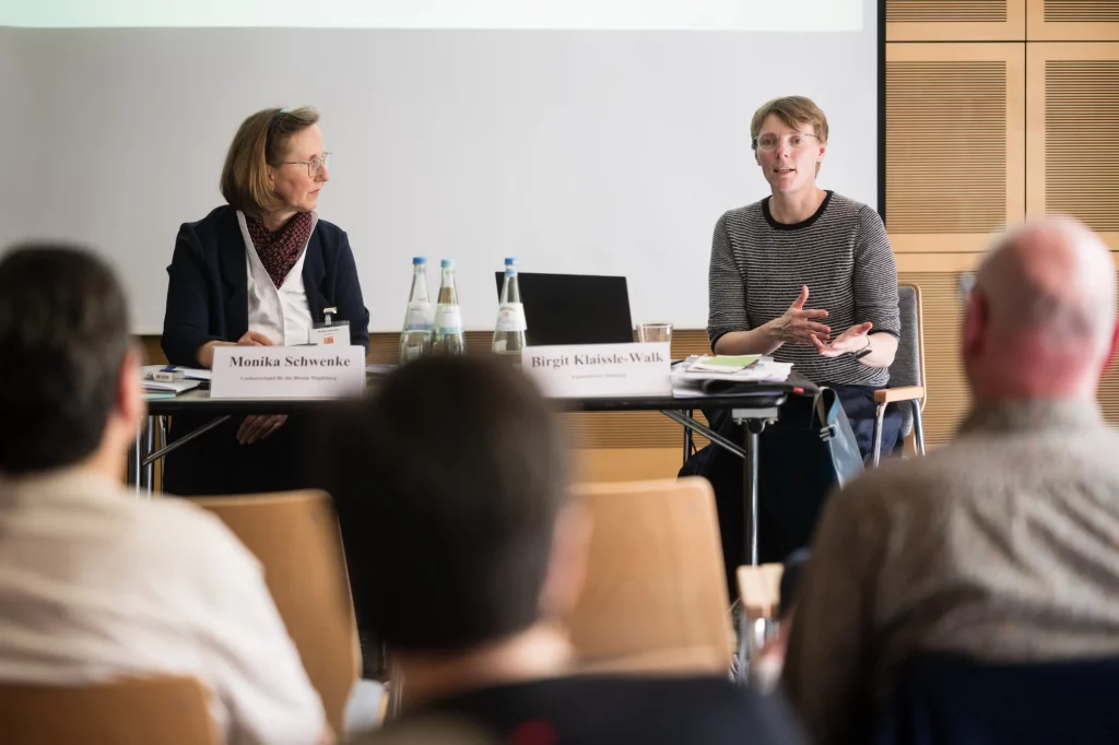 Zwei Frauen sitzen mit Namensschildern und Wasserflaschen an einem Tisch und sprechen während einer Präsentation oder Diskussion in einem Konferenzraum zu einem Publikum.