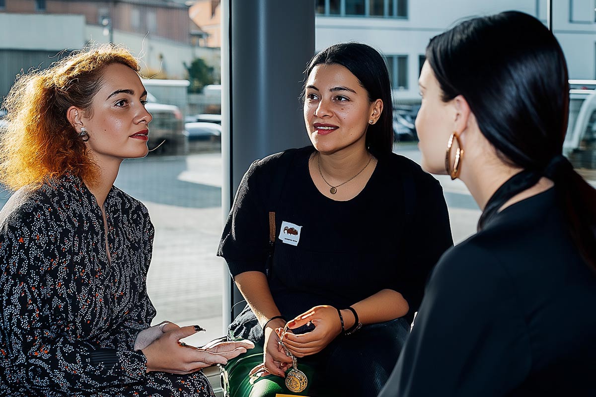 Drei Frauen sitzen drinnen und unterhalten sich. Eine hält ein Telefon, die andere trägt ein Namensschild. Durch das Fenster fällt Tageslicht ein.