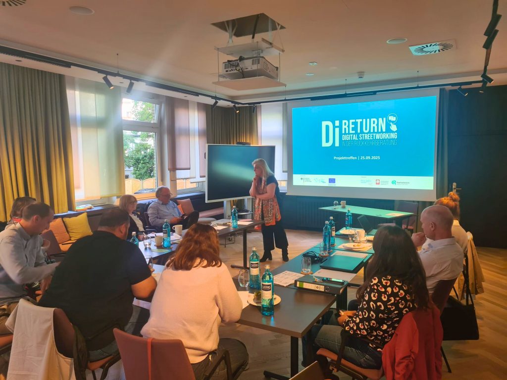 Eine Gruppe von Personen sitzt in einem Konferenzraum an Tischen und hört einem Vortragenden zu. Auf einem großen Bildschirm werden "Di RETURN Digitaler Pfahlbau" und Veranstaltungsdetails angezeigt.